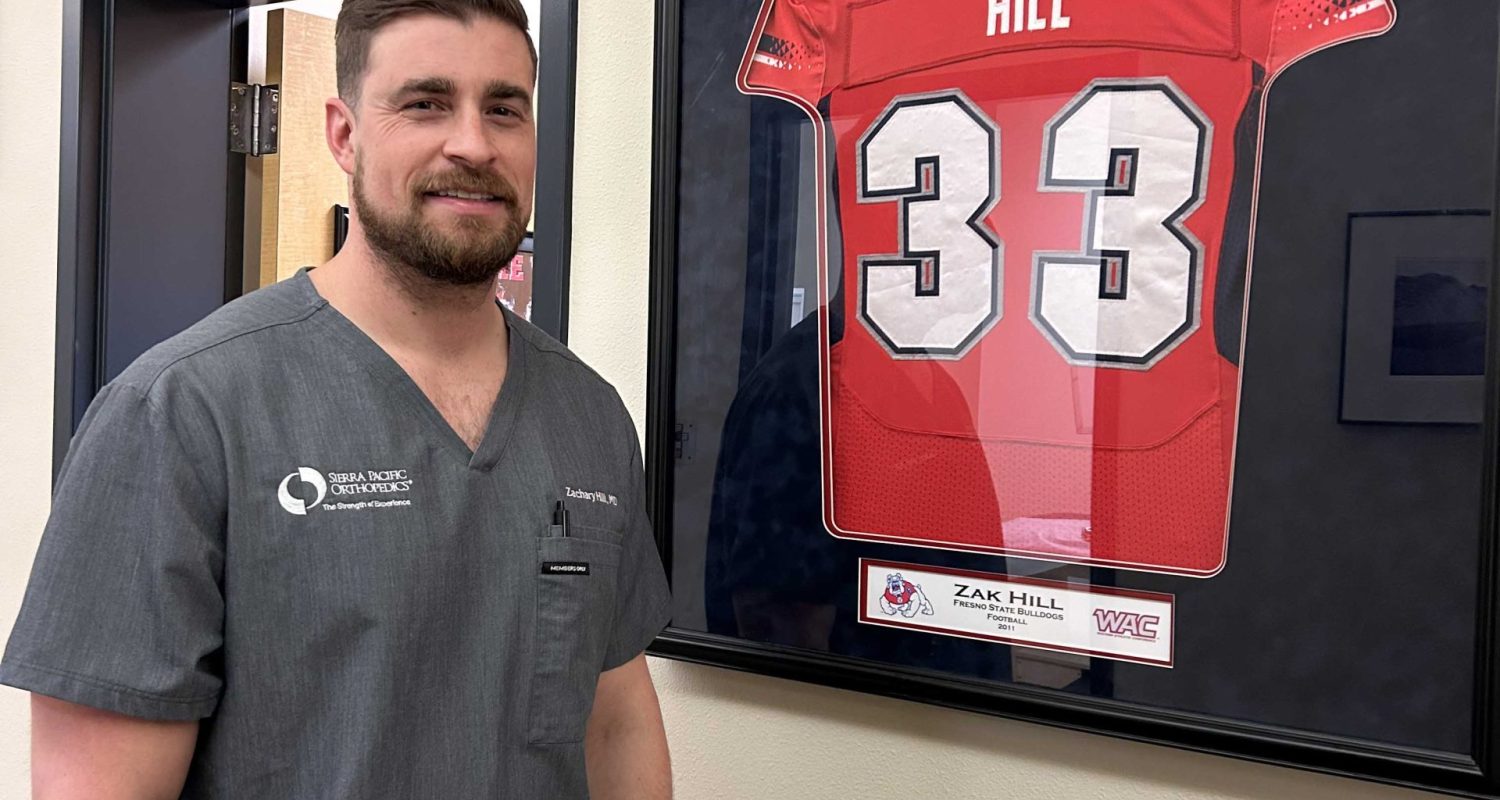 36661765-E208-429E-953A-B0E77BF6A0EC Zachary Hill ’17 at the Sierra Pacific Orthopedics office standing next to his framed Fresno State college football jersey.