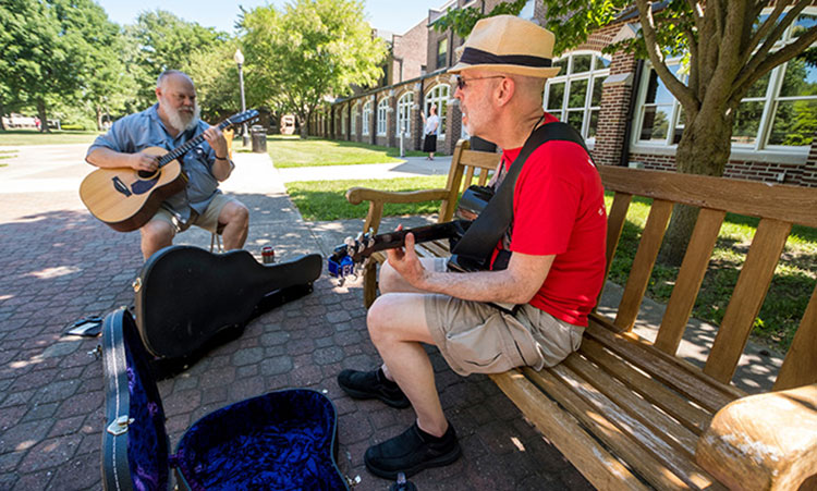 Two older alums play guitar in the shade outside of the dorms.