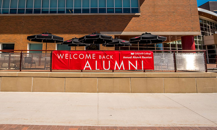 A red and white sign on the rails on the seating area of the JRC on the campus of Grinnell College. Text on the sign: Welcome Back Alumni - Annual Alumni Reunion.