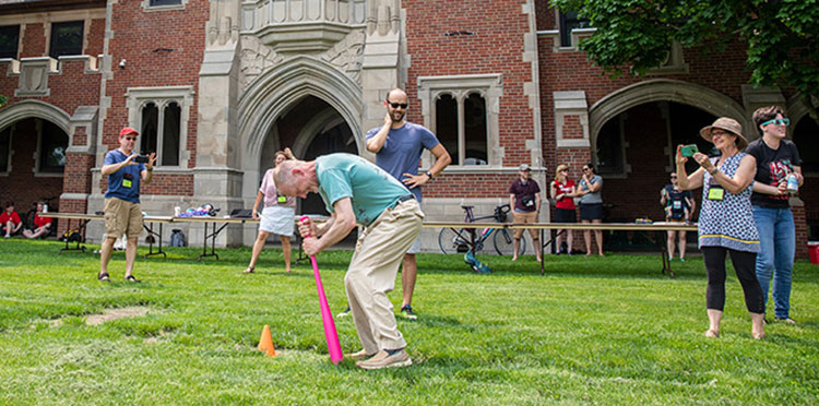 An alumnus spins on a pink wiffle bat during a relay race in front of Gates Tower.