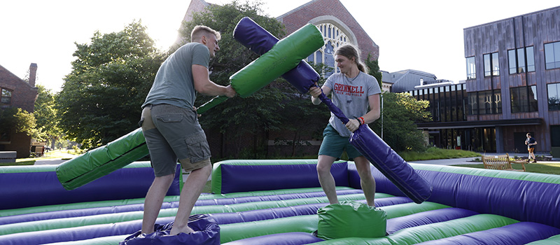 Two alums joust using foam jousting sticks in an inflatable jousting arena.