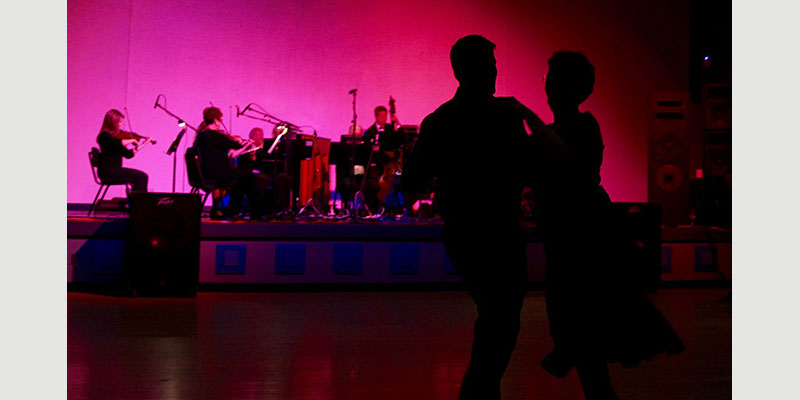 Two waltzing alumni are silhouetted against a purple and red backdrop. In the background, you can see a group of string musicians.